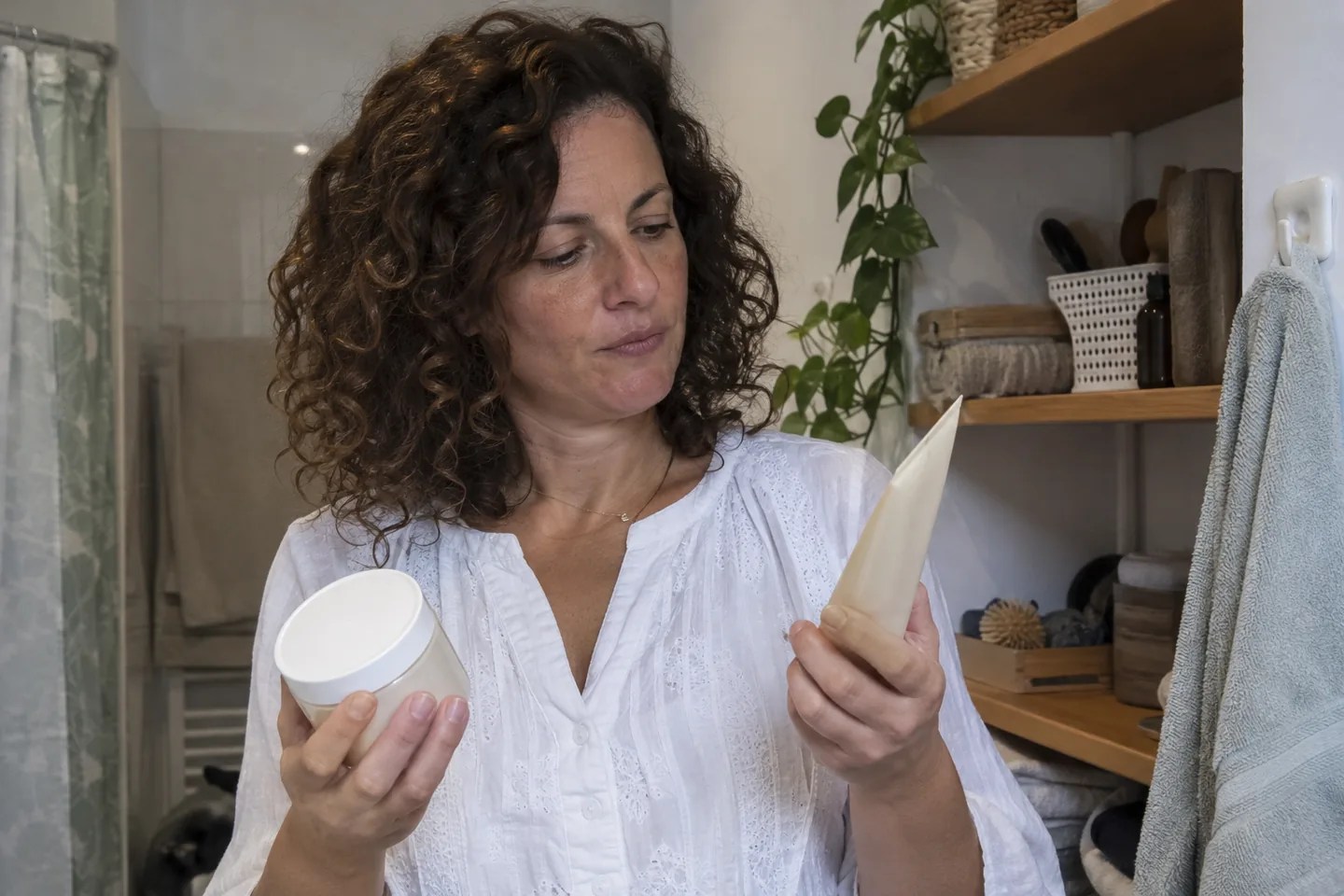 Femme aux cheveux bouclés tenant un tube clair et un pot blanc fermés.